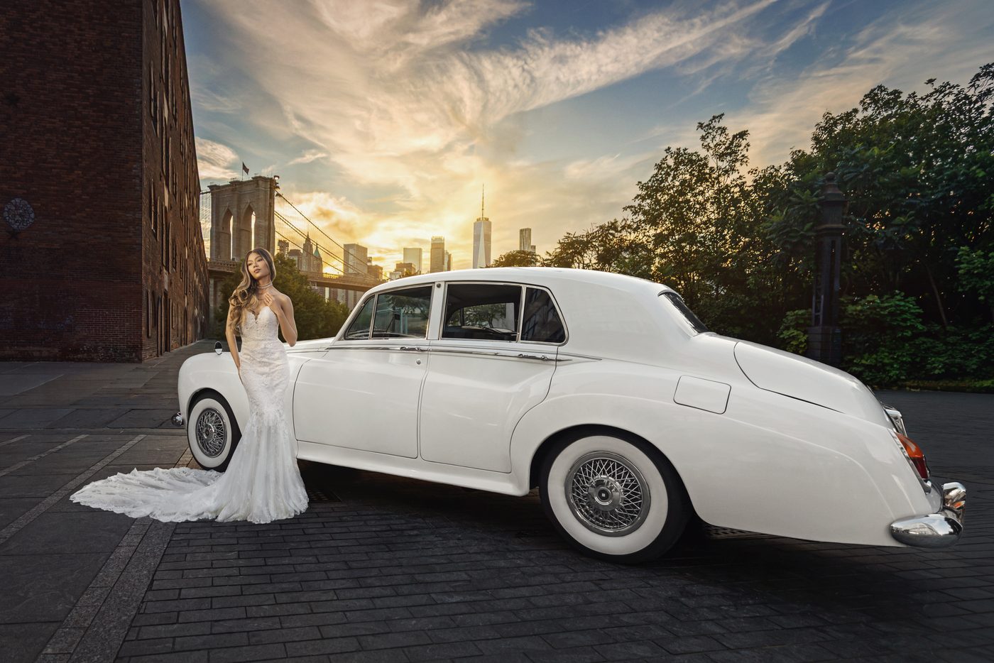 Bride in a lace gown beside a white vintage Rolls-Royce in DUMBO, Brooklyn, with the Brooklyn Bridge and Manhattan skyline at sunset